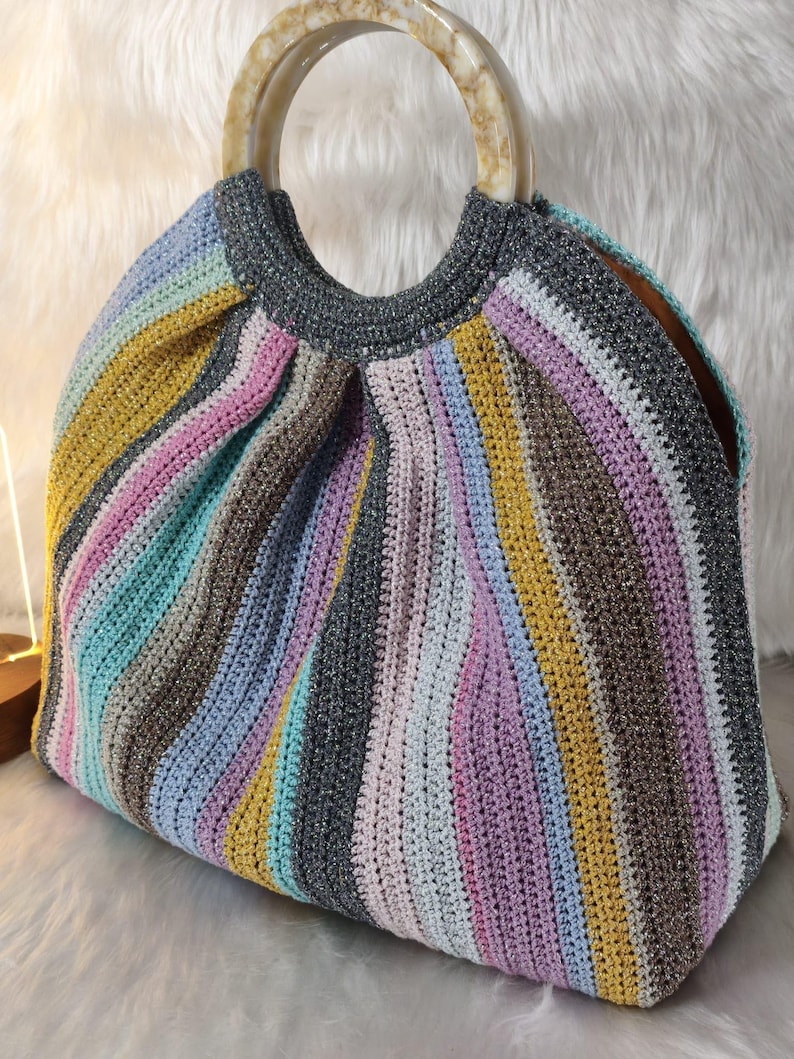 May include: A handmade crochet handbag with a striped pattern in various colors including yellow, pink, blue, and gray. The bag has a round, marbled handle and a structured shape, perfect for carrying essentials. The bag is sitting on a white, fluffy surface.