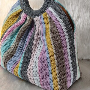 May include: A colorful, handmade crochet bag with a striped pattern in shades of pink, yellow, blue, gray, and brown. The bag has a rounded shape with a gray top and light-colored, circular handles.