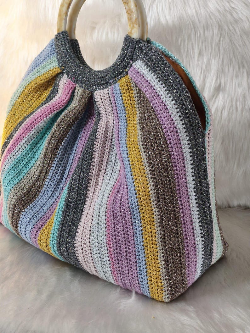 May include: A colorful, handmade crochet bag with a striped pattern in shades of pink, yellow, blue, gray, and brown. The bag has a rounded shape with a gray top and light-colored, circular handles.