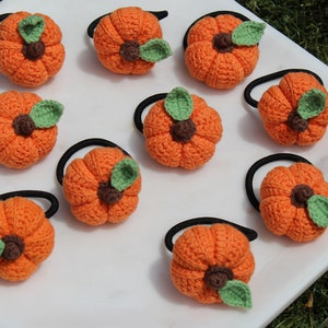 May include: Ten crocheted pumpkin hair ties in orange with green leaves. The hair ties are arranged on a white surface.