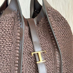 May include: A brown tote bag with a woven raffia body and leather accents. The bag features a leather strap with a gold-colored metal buckle. The handles are also made of brown leather.