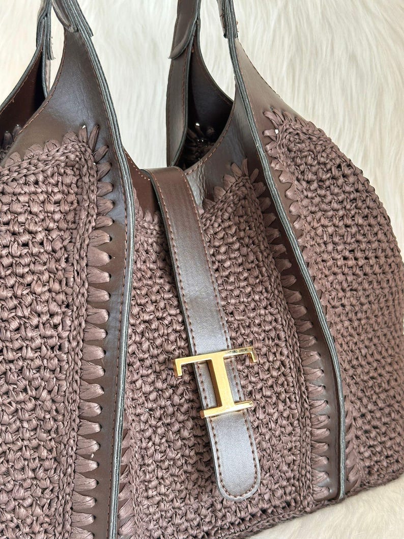 May include: A brown tote bag with a woven raffia body and leather accents. The bag features a leather strap with a gold-colored metal buckle. The handles are also made of brown leather.
