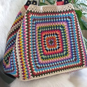May include: A large, colorful crochet tote bag with a granny square design. The bag features a vibrant mix of colors, including pink, blue, green, and yellow, with a cream-colored border and black leather handles. The bag is sitting on a white, fluffy surface.