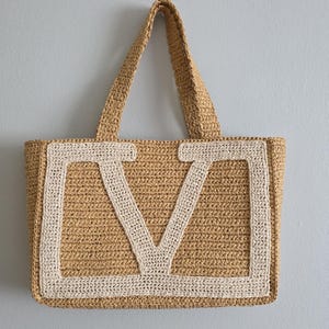 May include: A tan crocheted tote bag featuring two top handles and a large, cream-colored letter V stitched onto the front panel. The bag is constructed from a textured, woven material with a rectangular shape and a consistent, dense stitch pattern.