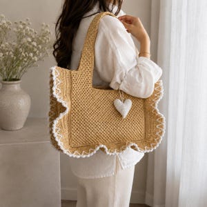 May include: A tan crochet tote bag featuring a rectangular body with wide, ruffled edges trimmed in white yarn. The bag includes two shoulder straps and a small, white, heart-shaped crochet charm hanging from the front of the main compartment.