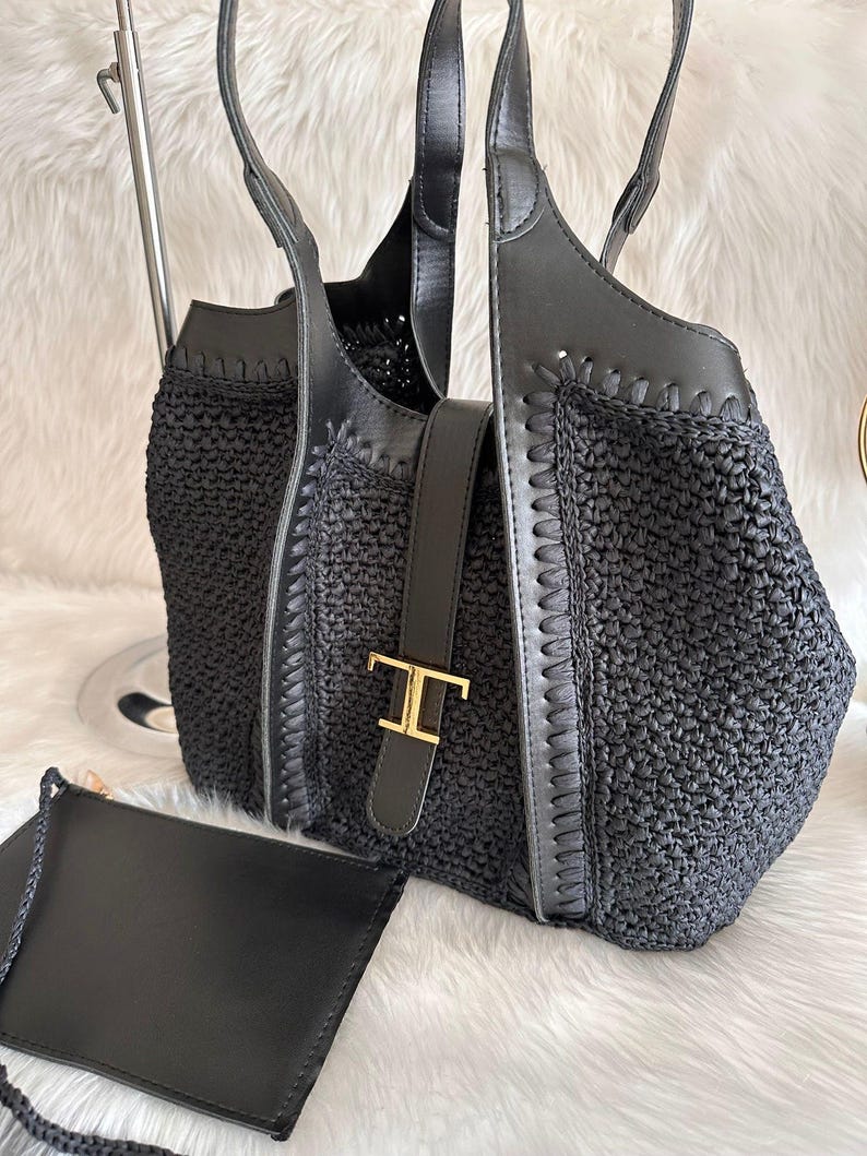 May include: Black woven tote bag with leather accents and a matching small pouch. The tote features a leather strap with a gold-tone buckle. The bag has two handles and a woven texture. The pouch has a braided strap.