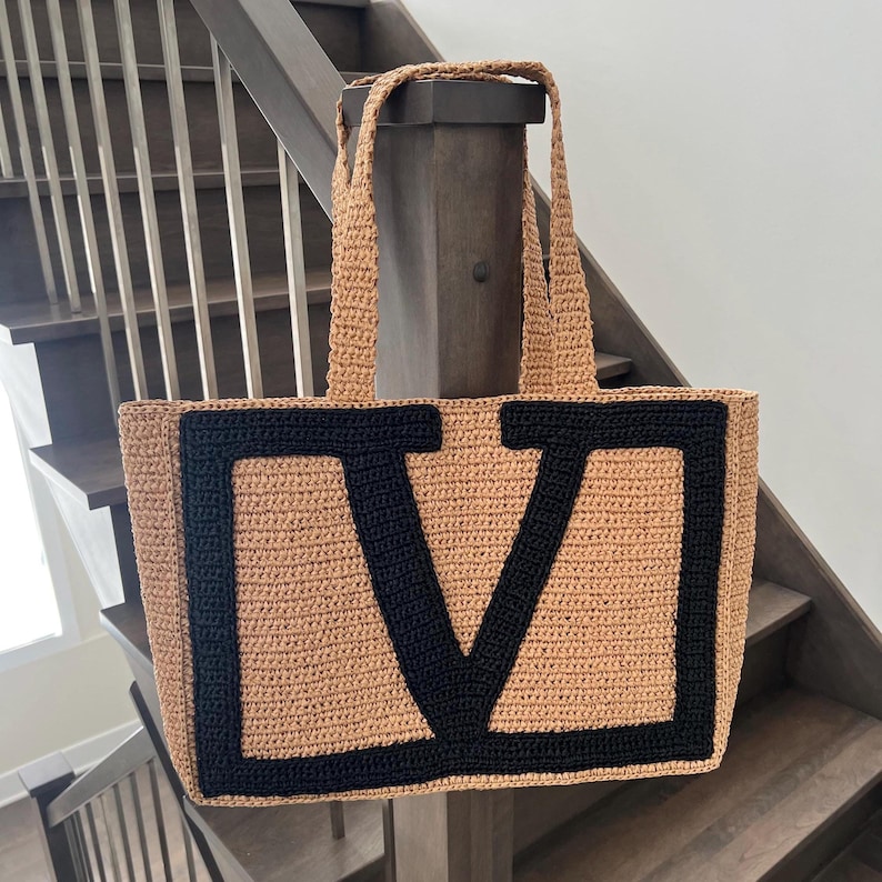 May include: A rectangular crochet tote bag in a tan color featuring a large, bold black letter V centered on the front. The bag has two matching tan crochet shoulder straps and a textured, woven appearance throughout the entire construction.