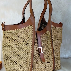 May include: A woven tote bag with brown leather accents. The bag features a light brown woven body, brown leather handles, and a buckle closure. The bag has a spacious interior and is suitable for everyday use.