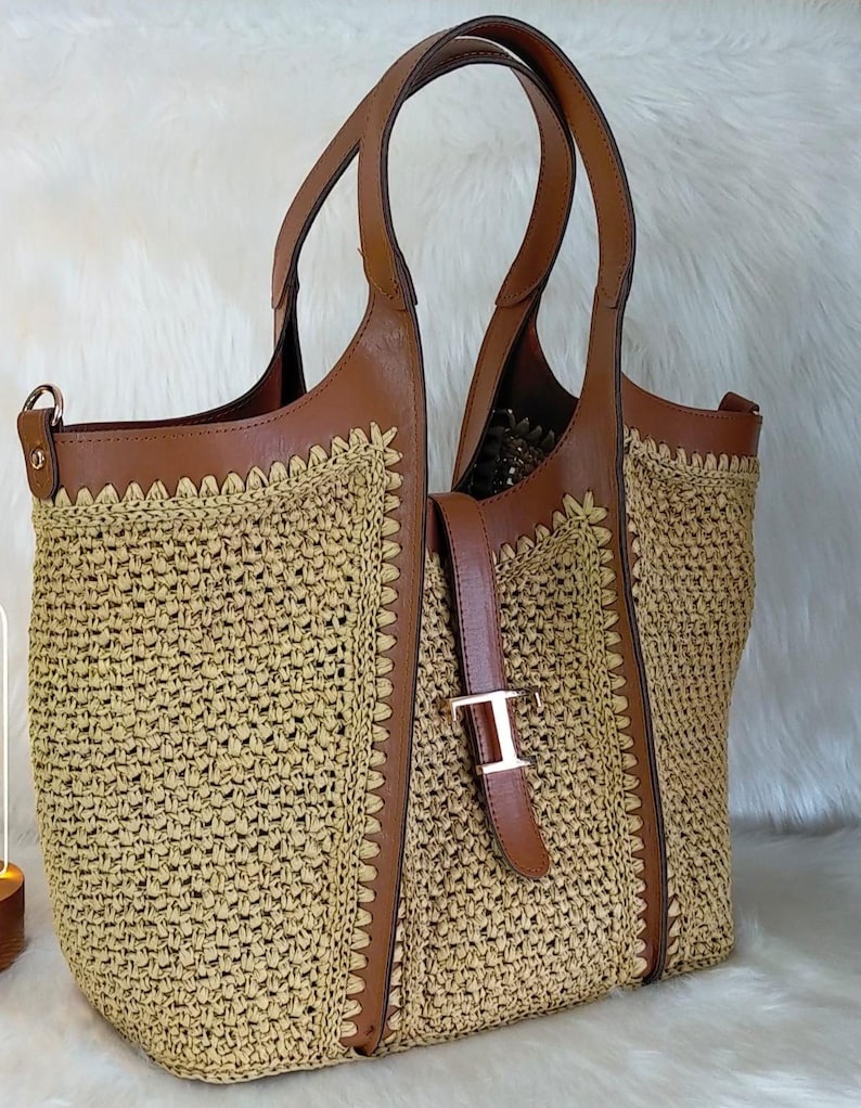 May include: A woven tote bag with brown leather accents. The bag features a light brown woven body, brown leather handles, and a buckle closure. The bag has a spacious interior and is suitable for everyday use.