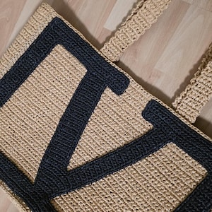 May include: A rectangular tote bag crafted from beige woven material featuring a large, dark charcoal gray letter V stitched onto the front surface. The bag includes two matching beige woven handles attached to the top edge of the structure.