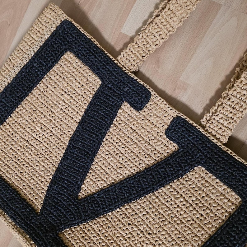 May include: A rectangular tote bag crafted from beige woven material featuring a large, dark charcoal gray letter V stitched onto the front surface. The bag includes two matching beige woven handles attached to the top edge of the structure.
