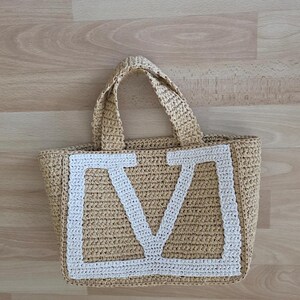 May include: A beige woven straw tote bag featuring two top handles and a large, white, stitched letter V centered on the front panel. The bag has a rectangular base and a textured, hand-crafted appearance with a consistent, repeating weave pattern.