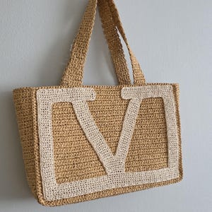 May include: A tan crochet tote bag featuring a large, cream-colored letter V appliqued on the front. The bag is constructed from a textured, woven material and includes two matching tan crochet shoulder straps attached to the top edge.