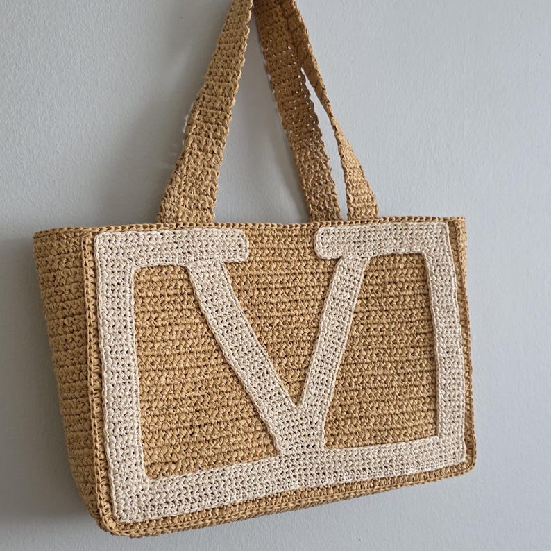 May include: A tan crochet tote bag featuring a large, cream-colored letter V appliqued on the front. The bag is constructed from a textured, woven material and includes two matching tan crochet shoulder straps attached to the top edge.