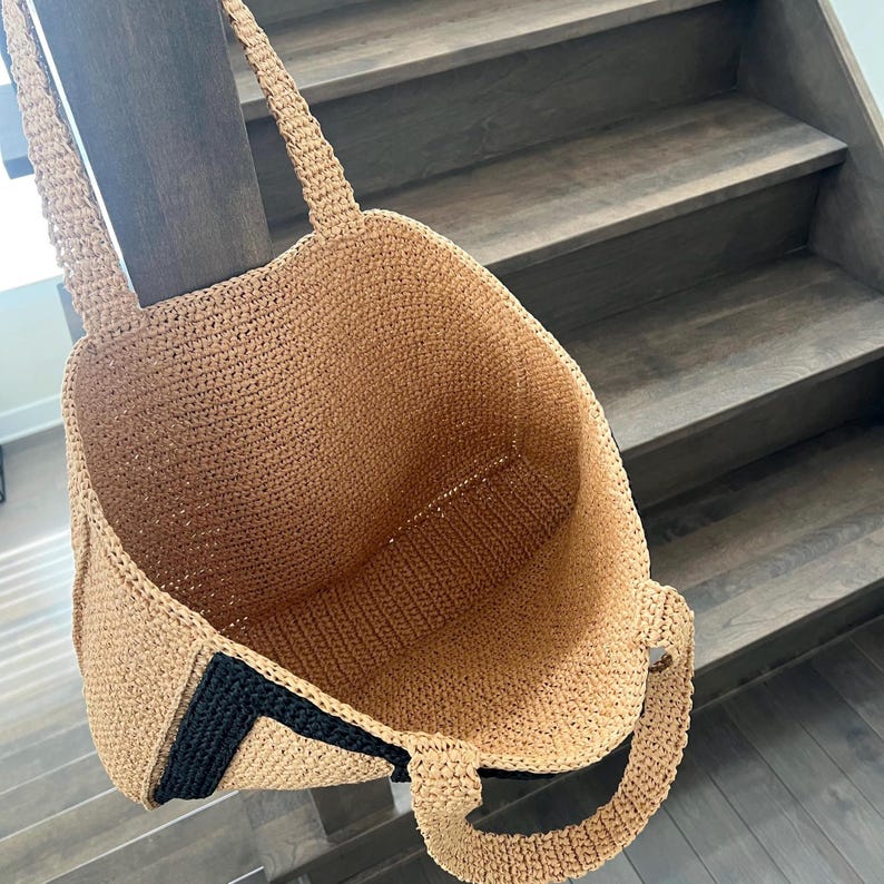 May include: A large, open-top tote bag crafted from tan crocheted material. The bag features two sturdy handles and a distinct black crocheted accent strip along the side panel. The interior is spacious and shows the consistent tan woven texture.