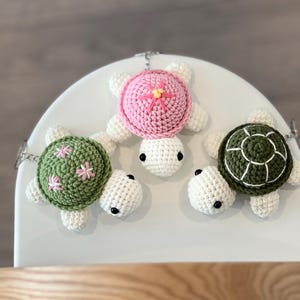 May include: Three handmade crochet turtle keyrings in pink, green, and white. The turtles have black eyes and floral patterns. The keyrings are displayed on a white surface.