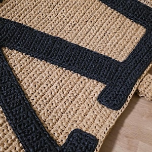 May include: A hand-crocheted bag featuring a natural tan raffia-like base with a bold, black letter A stitched onto the front. The texture is consistent throughout, showcasing a dense, woven pattern that creates a structured, durable appearance.