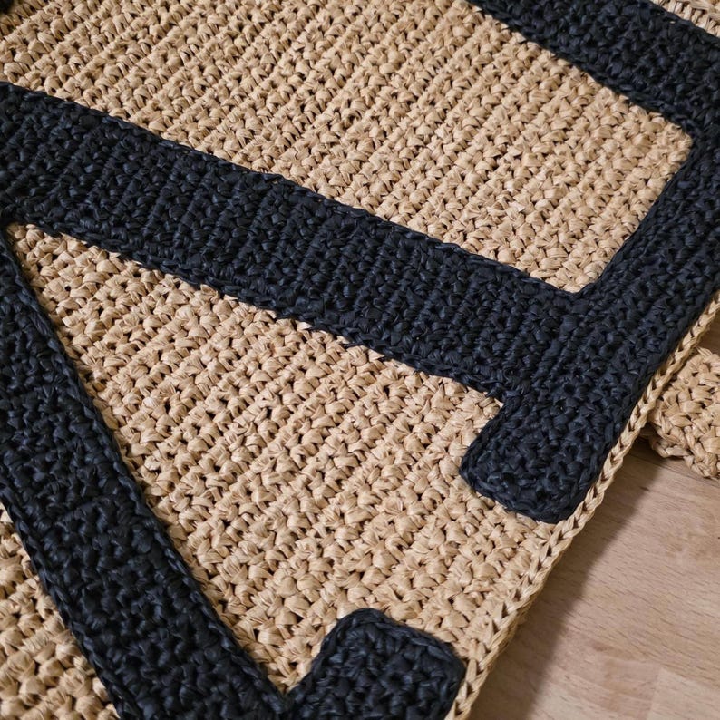 May include: A hand-crocheted bag featuring a natural tan raffia-like base with a bold, black letter A stitched onto the front. The texture is consistent throughout, showcasing a dense, woven pattern that creates a structured, durable appearance.