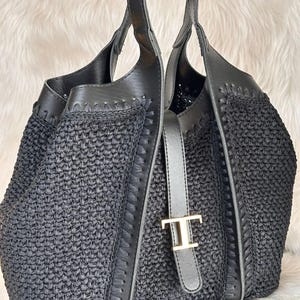 May include: Black woven tote bag with leather accents and handles. The bag features a silver-toned buckle with a gold-colored 'T' emblem. The bag's design includes a woven texture and stitched leather trim, creating a stylish accessory.