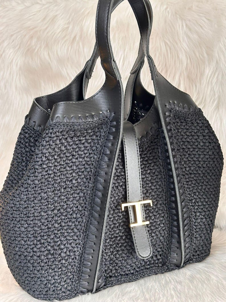 May include: Black woven tote bag with leather accents and handles. The bag features a silver-toned buckle with a gold-colored 'T' emblem. The bag's design includes a woven texture and stitched leather trim, creating a stylish accessory.