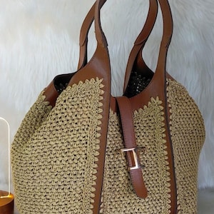 May include: A woven straw tote bag with brown leather accents. The bag features a woven body, brown leather handles, and a buckle closure. The bag is a light tan color with brown leather trim and a gold-colored buckle.