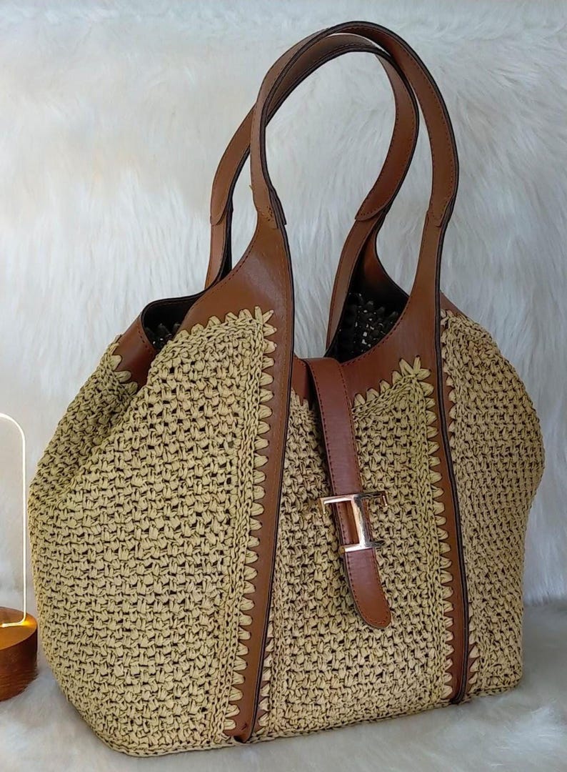 May include: A woven straw tote bag with brown leather accents. The bag features a woven body, brown leather handles, and a buckle closure. The bag is a light tan color with brown leather trim and a gold-colored buckle.