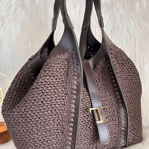May include: A brown woven tote bag with leather accents. The bag features a woven body, leather handles, and a leather strap with a gold-colored buckle. The bag is designed with a spacious interior and a stylish, textured appearance.