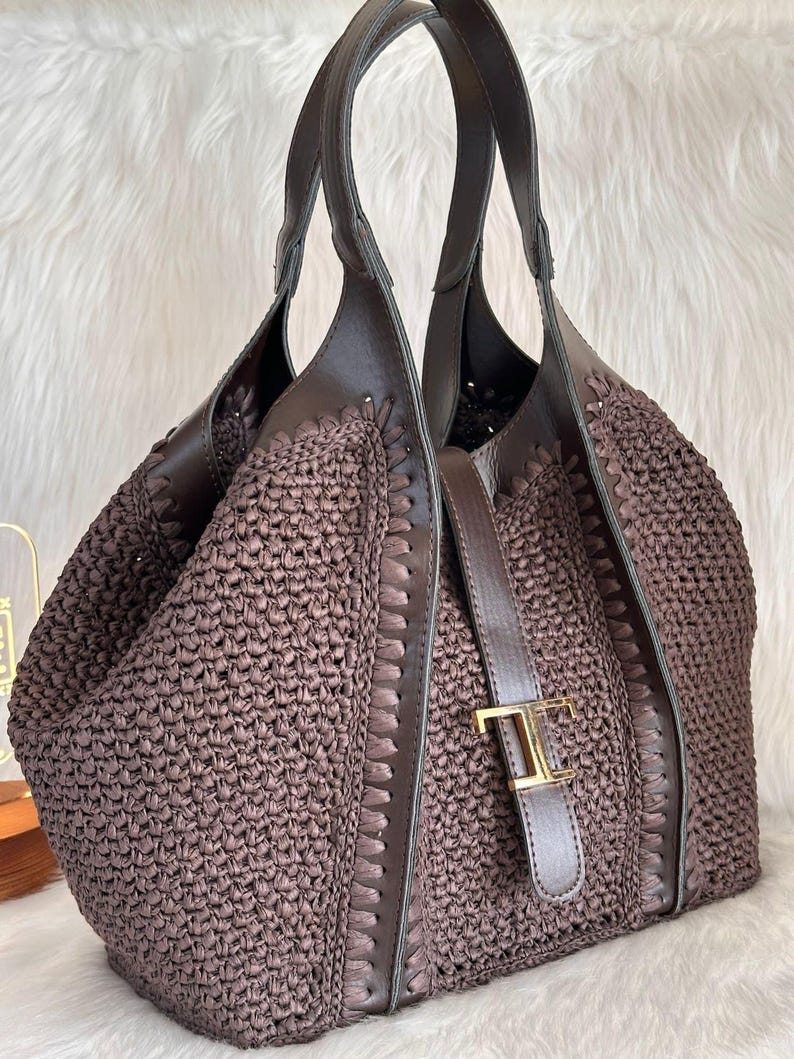May include: A brown woven tote bag with leather accents. The bag features a woven body, leather handles, and a leather strap with a gold-colored buckle. The bag is designed with a spacious interior and a stylish, textured appearance.