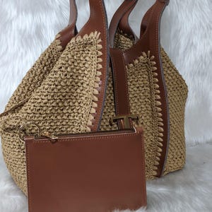 May include: A woven straw tote bag with brown leather accents and handles. A matching brown leather zip-up pouch with a gold zipper is in front of the bag. The bag has a light brown color and is set against a white background.