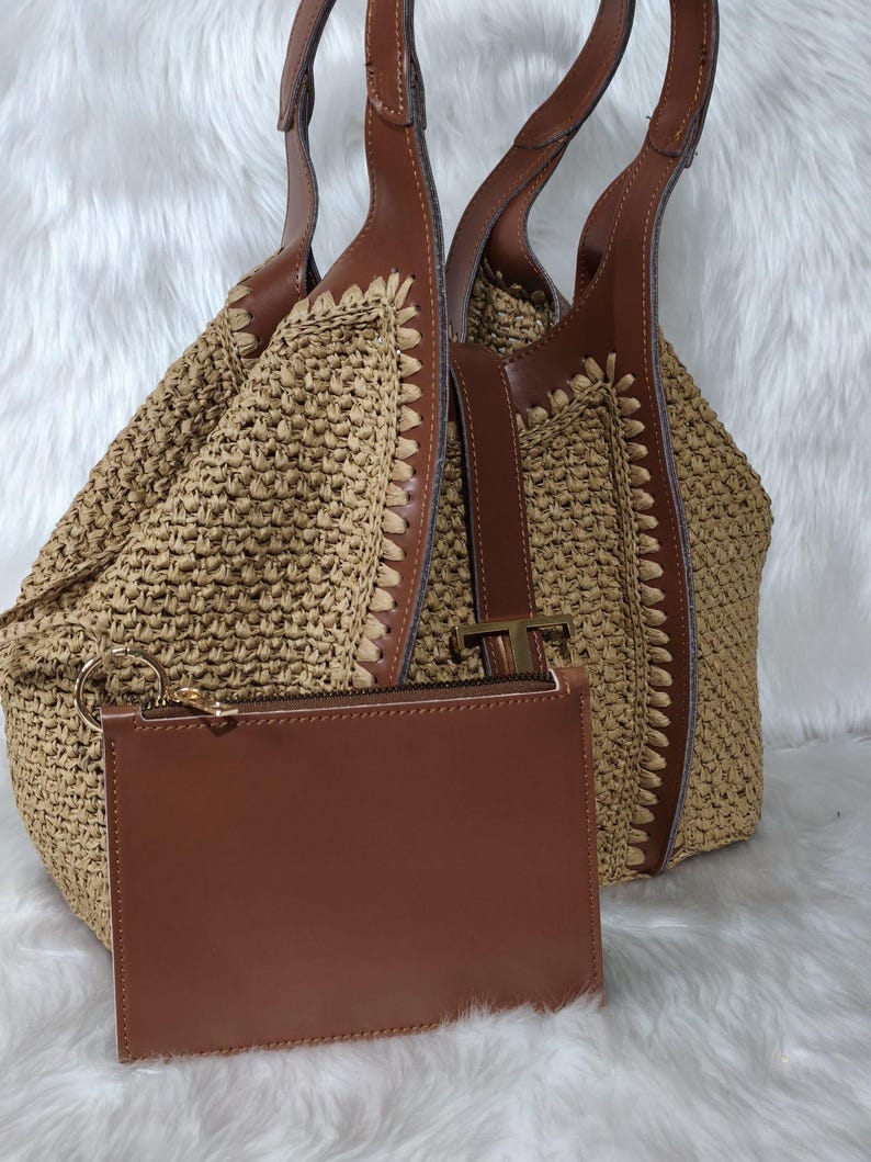 May include: A woven straw tote bag with brown leather accents and handles. A matching brown leather zip-up pouch with a gold zipper is in front of the bag. The bag has a light brown color and is set against a white background.