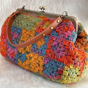 May include: A colorful, handmade crochet handbag with a brown leather handle and a metal clasp. The bag features a patchwork design in shades of orange, blue, green, and pink. The handle is secured with small gold-colored rivets.