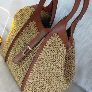 May include: A woven straw tote bag with brown leather accents. The bag features a buckle closure and two brown leather handles. The bag is a light tan color with a woven pattern and brown leather trim.