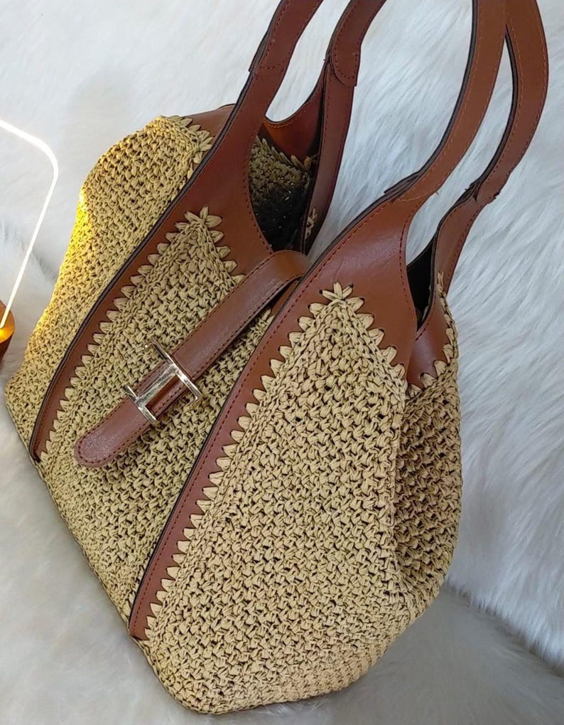 May include: A woven straw tote bag with brown leather accents. The bag features a buckle closure and two brown leather handles. The bag is a light tan color with a woven pattern and brown leather trim.