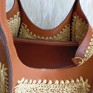 May include: A brown and beige tote bag with a woven straw design. The bag features brown leather straps and trim, with a zippered interior. The bag's interior is a light brown color, and the bag has a gold-colored metal ring.