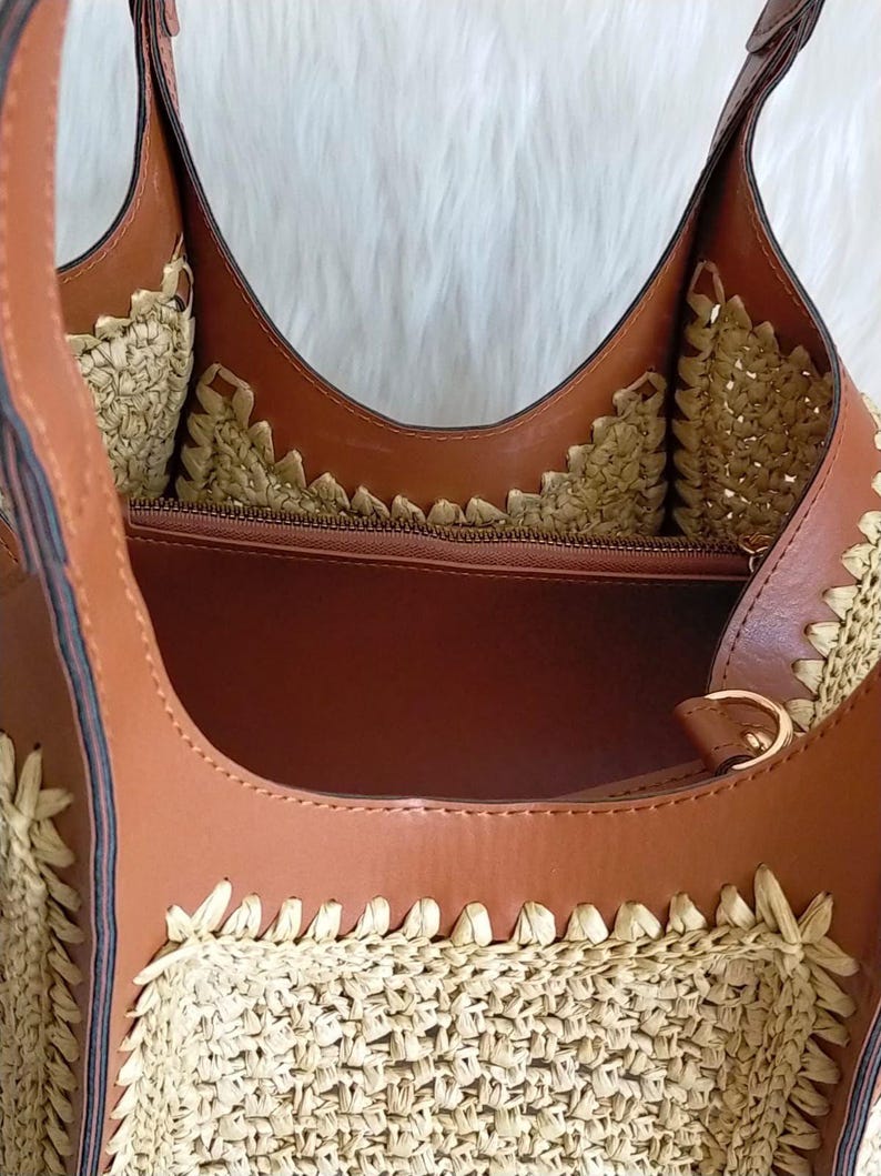 May include: A brown and beige tote bag with a woven straw design. The bag features brown leather straps and trim, with a zippered interior. The bag's interior is a light brown color, and the bag has a gold-colored metal ring.