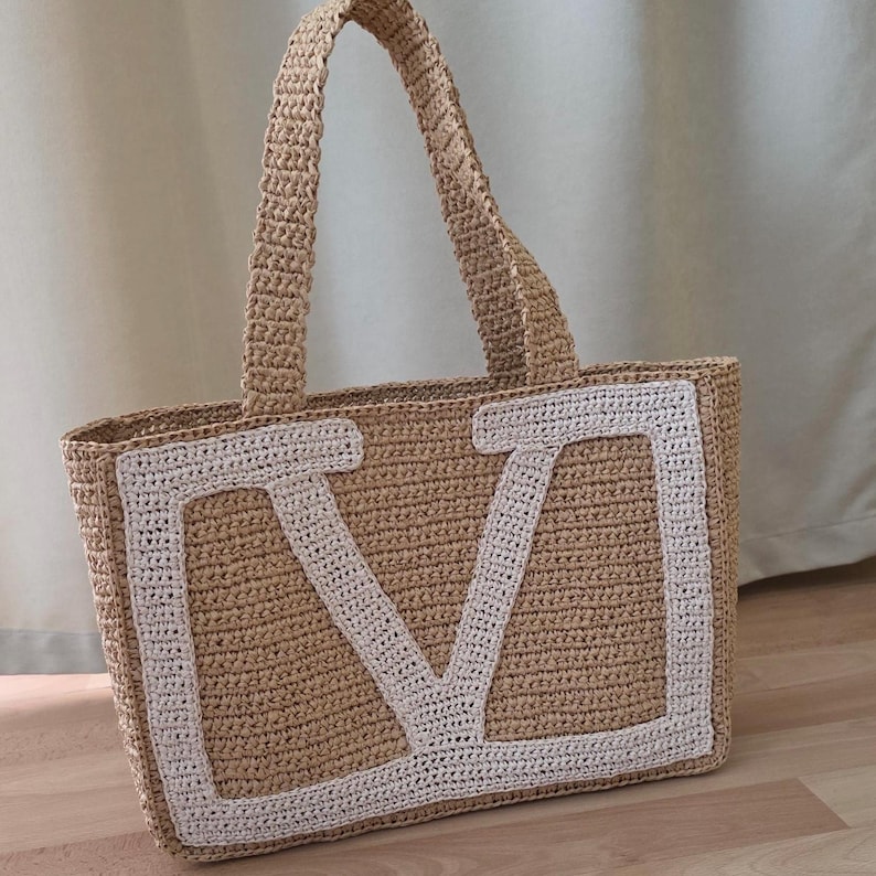 May include: A rectangular tote bag crafted from tan crocheted material, featuring two matching top handles. The front of the bag displays a large, white, block-letter V stitched onto the center, creating a distinct contrast against the tan base.