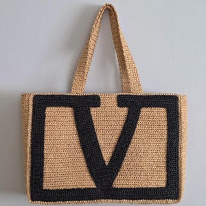 May include: A rectangular tote bag crafted from tan woven straw material, featuring two matching top handles. The front of the bag displays a large, bold letter V stitched in thick black yarn, creating a high-contrast design against the natural tan background.