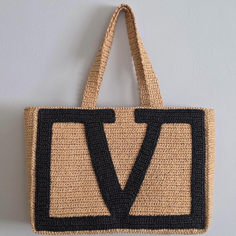 May include: A rectangular tote bag crafted from tan woven straw material, featuring two matching top handles. The front of the bag displays a large, bold letter V stitched in thick black yarn, creating a high-contrast design against the natural tan background.