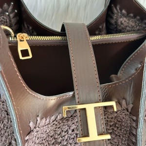 May include: A brown handbag with a woven texture and leather accents. The bag features a gold-colored clasp and a leather strap with a gold-colored letter "T" buckle. The interior of the bag is visible, showcasing a dark brown lining.