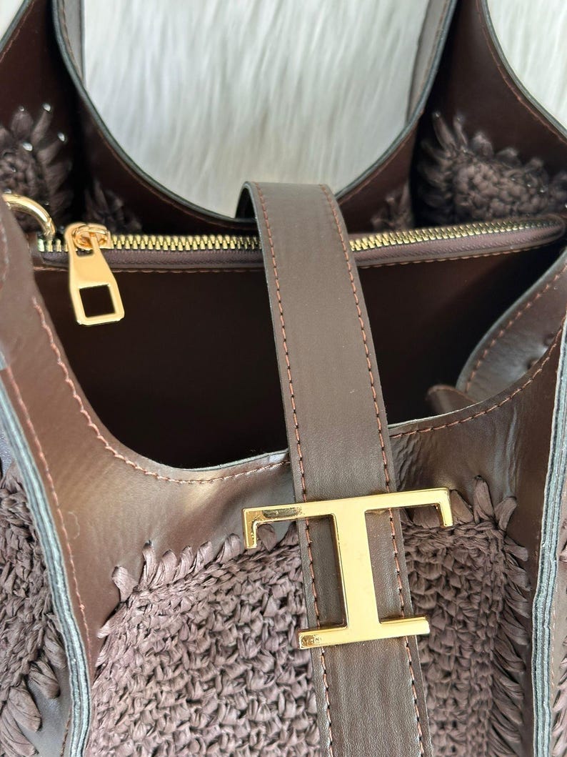 May include: A brown handbag with a woven texture and leather accents. The bag features a gold-colored clasp and a leather strap with a gold-colored letter "T" buckle. The interior of the bag is visible, showcasing a dark brown lining.