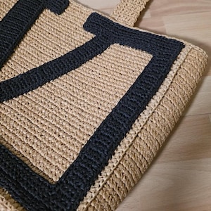 May include: A beige woven straw tote bag featuring a large, bold black letter U stitched onto the front surface. The bag is constructed with a textured, braided material and includes a matching woven handle extending from the top edge.