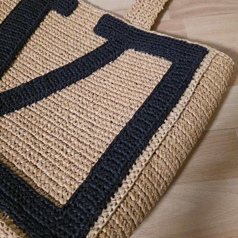 May include: A beige woven straw tote bag featuring a large, bold black letter U stitched onto the front surface. The bag is constructed with a textured, braided material and includes a matching woven handle extending from the top edge.