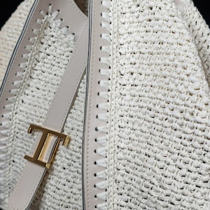 May include: Close-up of a cream-colored woven handbag with a gold-tone metal clasp. The bag features light beige leather trim and stitching details. The woven material has a textured appearance.