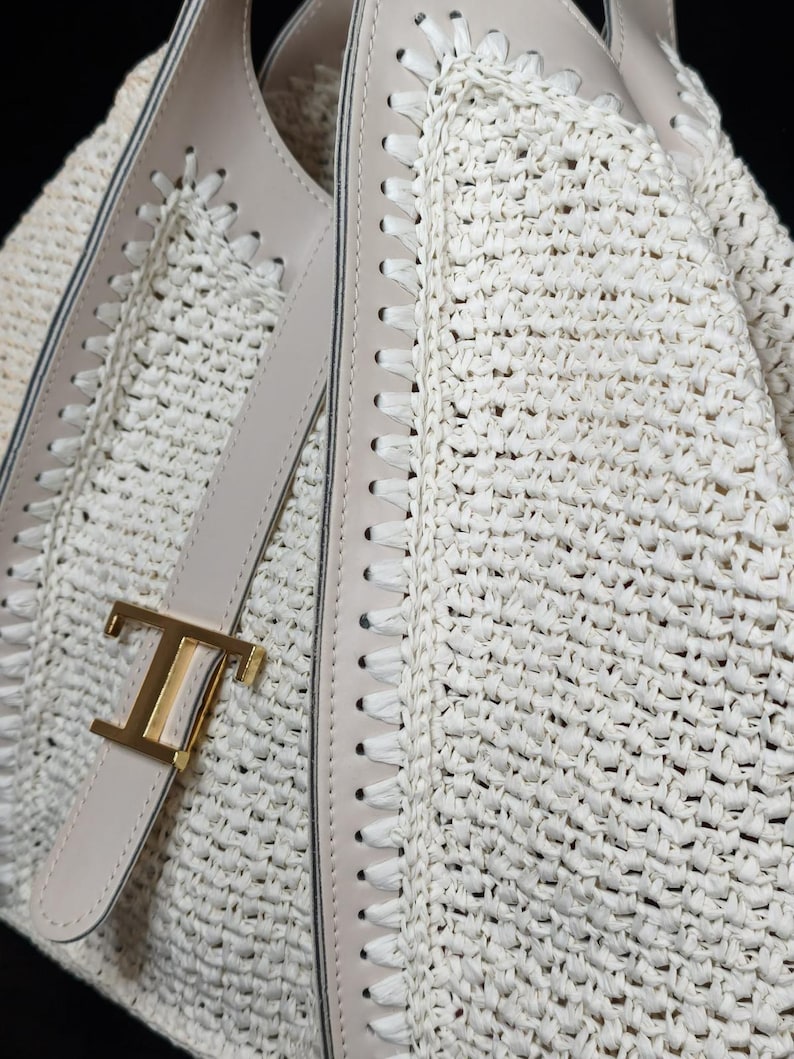 May include: Close-up of a cream-colored woven handbag with a gold-tone metal clasp. The bag features light beige leather trim and stitching details. The woven material has a textured appearance.