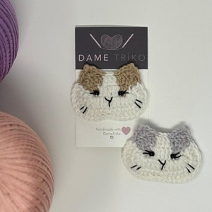 Op de afbeelding: Twee gehaakte kattengezichten, een met bruin en wit bont en de andere met grijs en wit bont. De kattengezichten zijn handgemaakt en hebben een schattige, cartoonachtige uitdrukking. De tekst "DAME TRIKO" is zichtbaar op een kaart achter de kattengezichten.