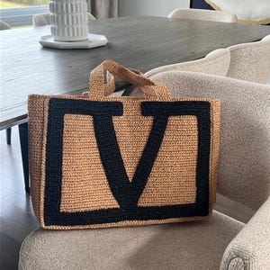 May include: A rectangular tote bag crafted from tan woven material, featuring two short top handles and a large, bold black letter V stitched prominently onto the front panel. The bag has a structured shape with a flat base and a textured finish.