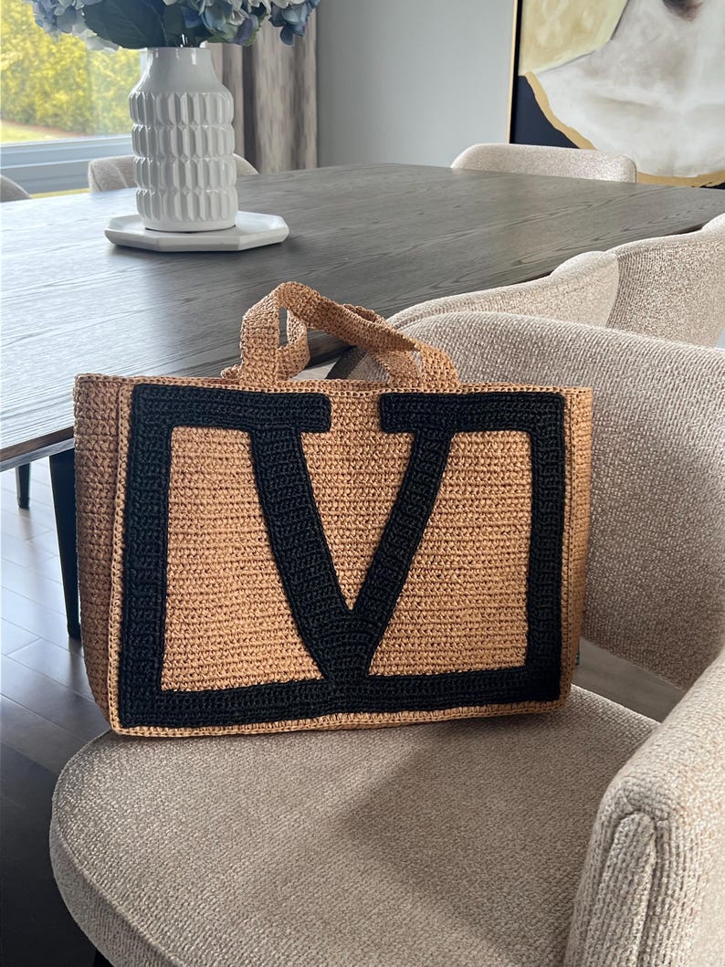 May include: A rectangular tote bag crafted from tan woven material, featuring two short top handles and a large, bold black letter V stitched prominently onto the front panel. The bag has a structured shape with a flat base and a textured finish.
