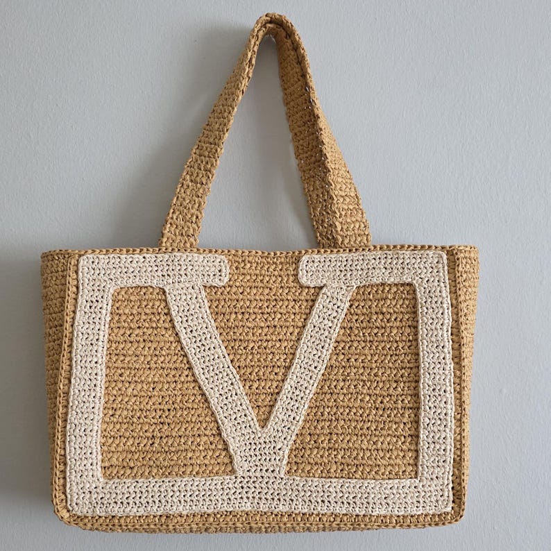 May include: A tan crocheted tote bag featuring a large, cream-colored letter V stitched onto the front panel. The bag has two matching tan crocheted handles and a rectangular body constructed from a textured, woven-style yarn material.