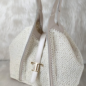 May include: Off-white woven tote bag with beige leather accents and handles. The bag features a gold-tone buckle and a structured design. The bag is made of a woven material and has a spacious interior, perfect for everyday use.