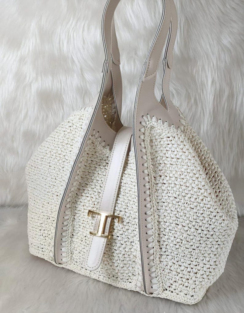 May include: Off-white woven tote bag with beige leather accents and handles. The bag features a gold-tone buckle and a structured design. The bag is made of a woven material and has a spacious interior, perfect for everyday use.
